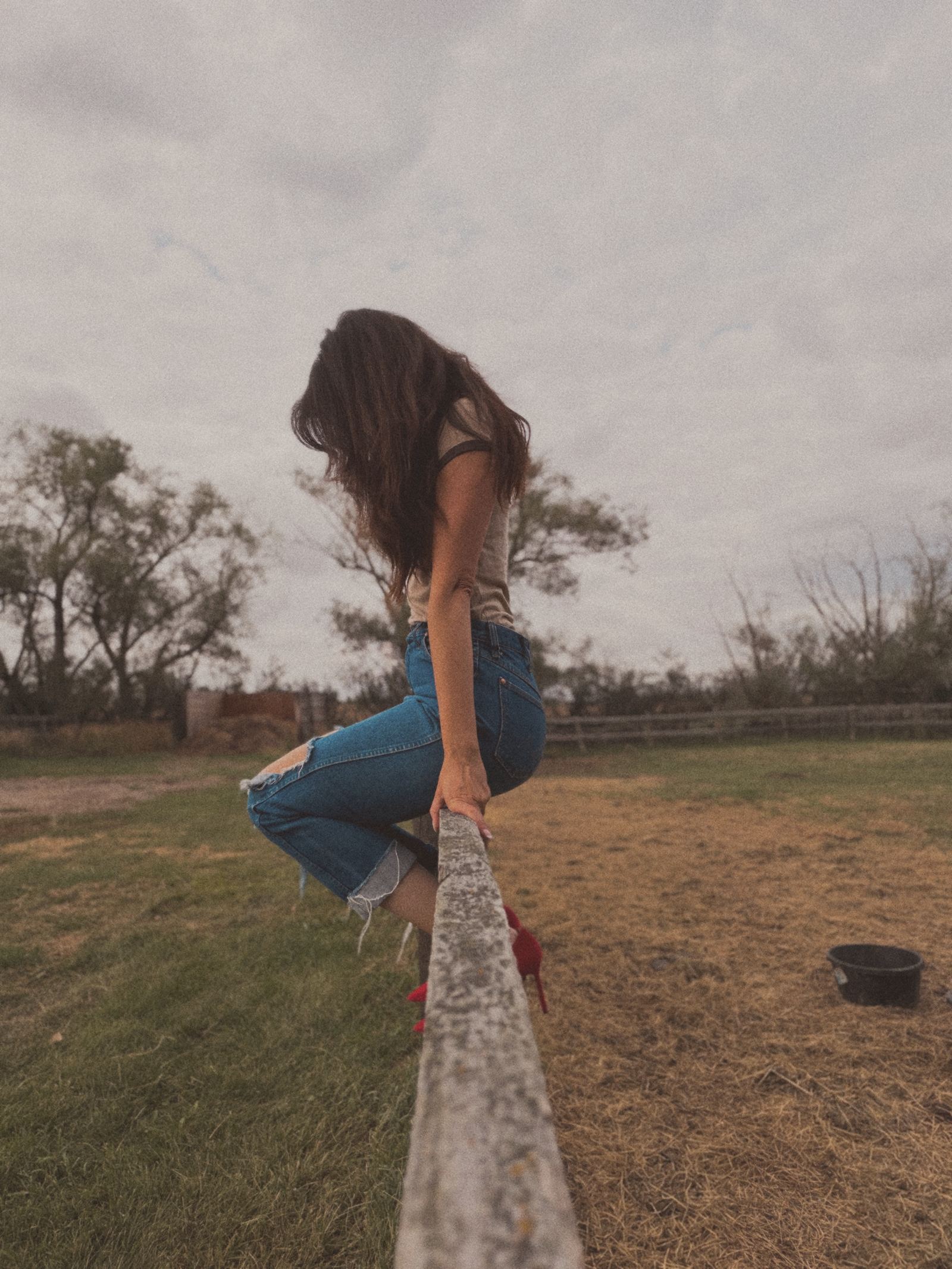 Sandra balancing on a weathered wooden fence rail in a grassy paddock under a cloudy sky, in jeans and red heels.